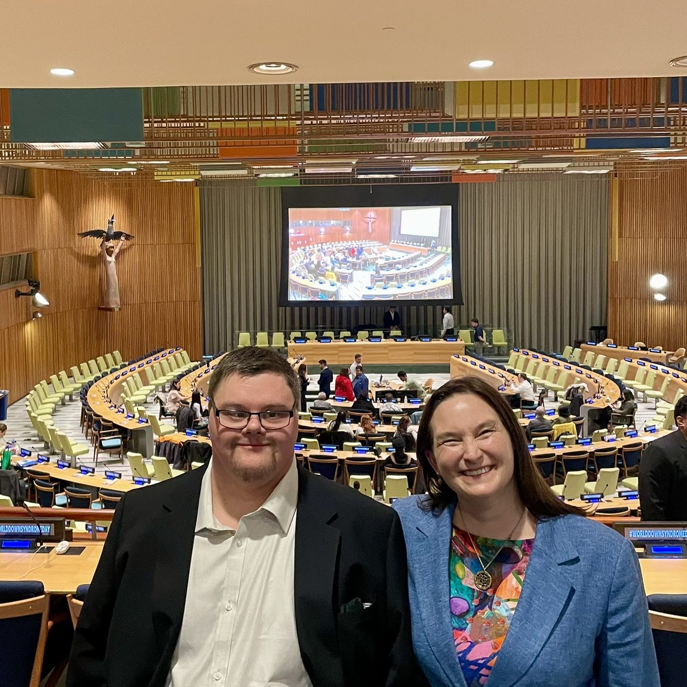 Tim and Kathy standing in front of the Trusteeship Council Chambers at the United Nations Headquarters Building in New York. Behind Tim and Kathy are people setting up for the session.