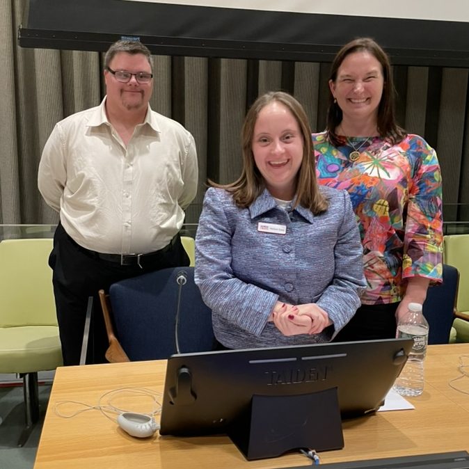 A photo of Tim Cologon, Madison Essig, and Kathy Cologon standing together behind the desk at the front of the Trusteeship Council Chambers in the United Nations Headquarters, New York