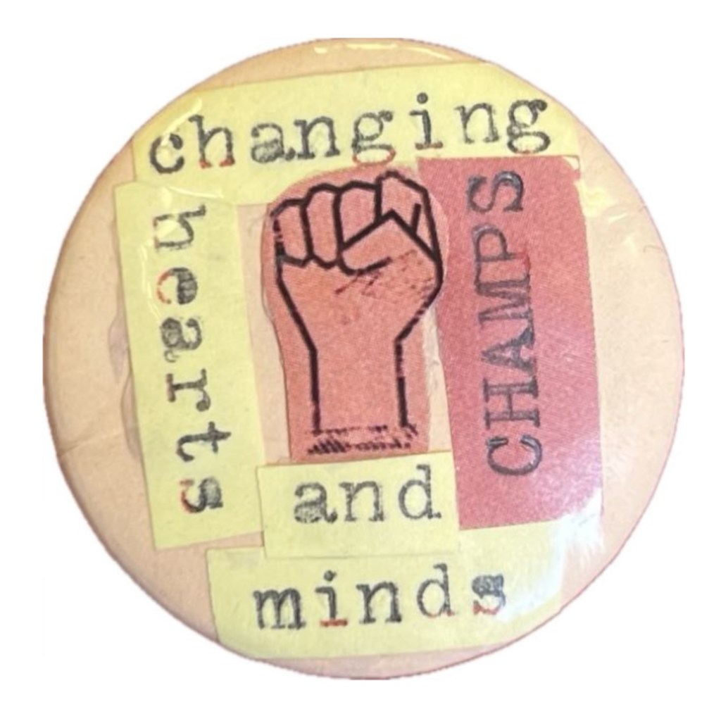 A photo of a pin on badge that has the words 'Changing hearts and minds, CHAMPS' around a fist representing strength and solidarity.