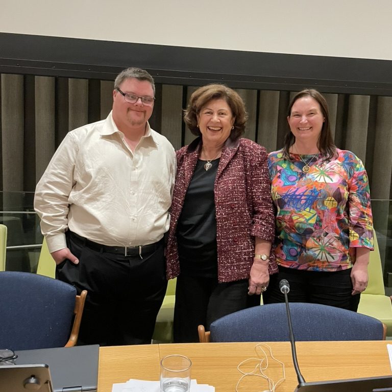 A photo of Tim Cologon, Bridget Sneddon, and Kathy Cologon standing together behind the desk at the front of the Trusteeship Council Chambers in the United Nations Headquarters, New York