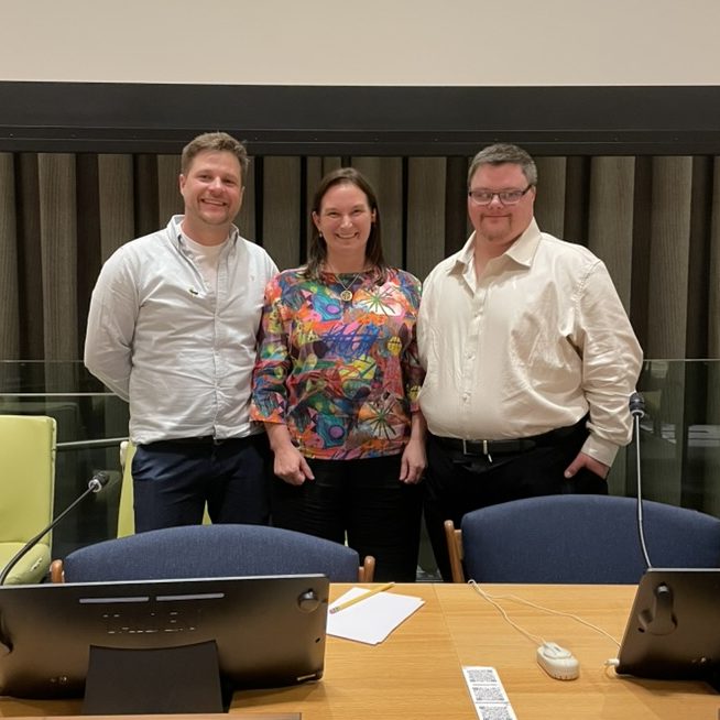 A photo of Robin Gibson, Kathy Cologon, and Tim Cologon standing together behind the desk at the front of the Trusteeship Council Chambers in the United Nations Headquarters, New York