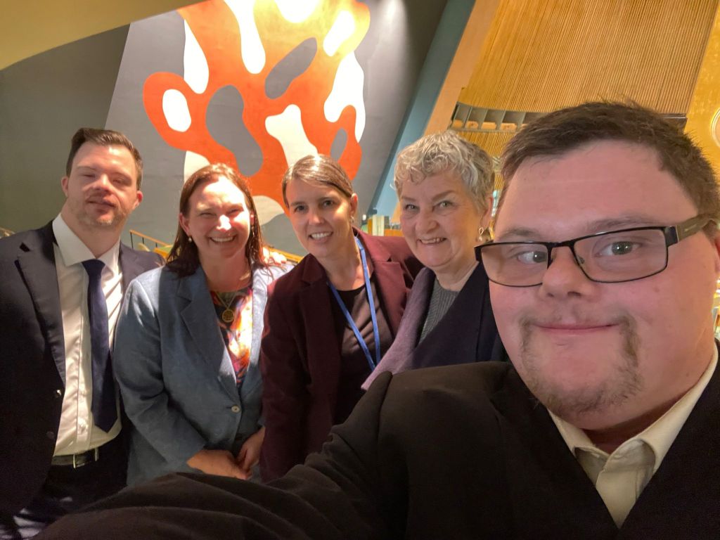 A photo of Michael Cox, Kathy Cologon, Sheona McKenna, Nikki Cox, and Tim Cologon standing together in front of a Fernand Leger Mural in the General Assembly at the United Nations Headquarters, New York