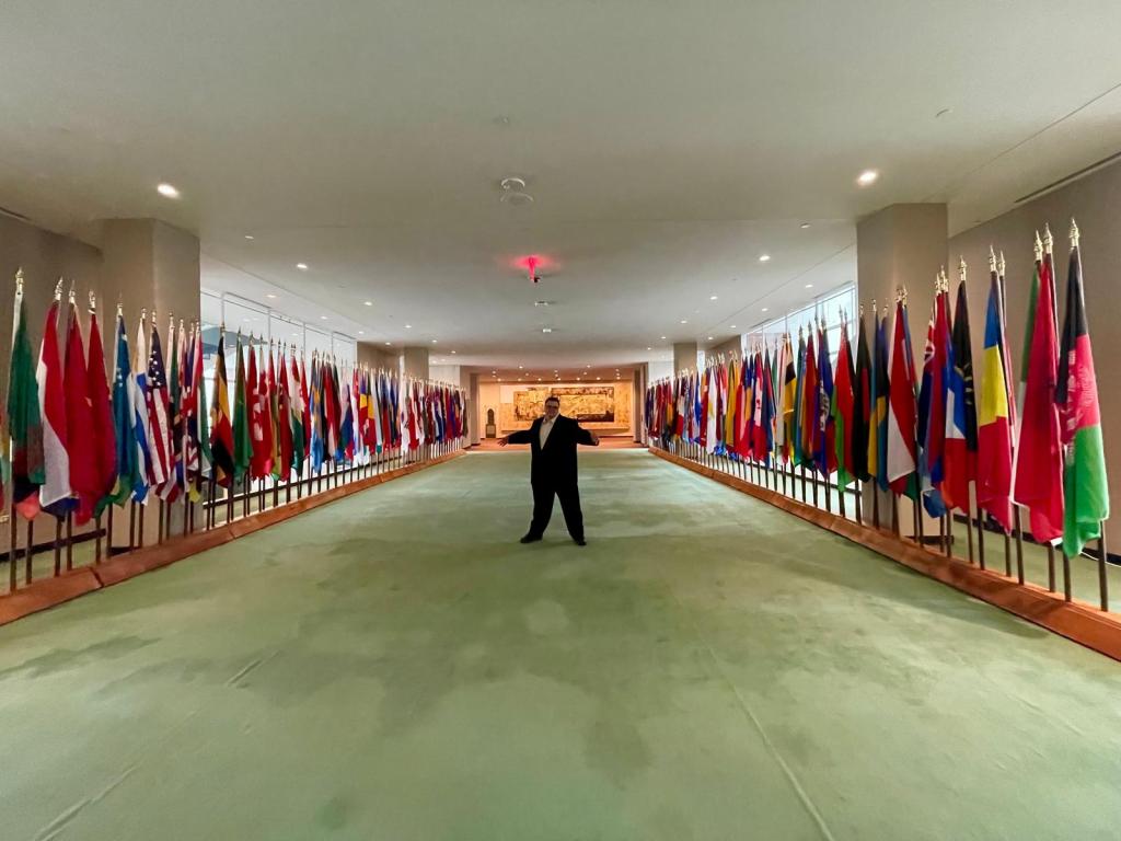 A photo of Tim Cologon standing in the middle of the display of the flags of all the member countries within the United Nations building.