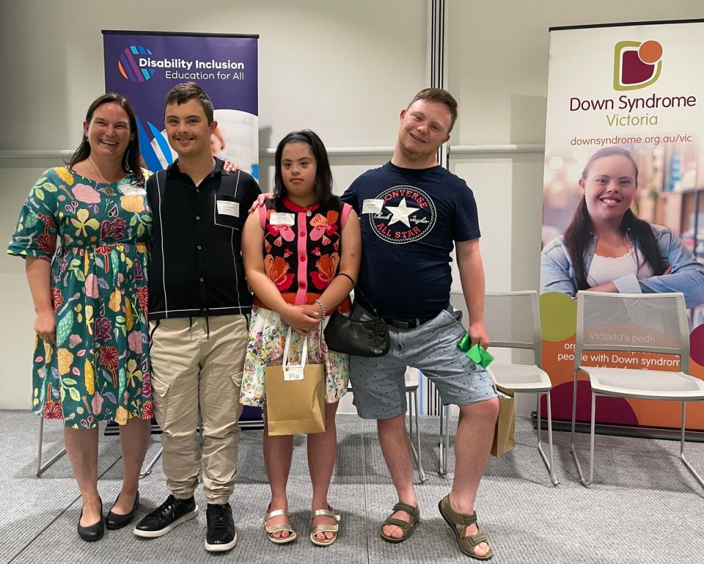 A photo of Kathy Cologon, Harry Sencek, Pia Bolger, and Owen Roberts standing on the conference stage in front of a poster for Down Syndrome Victoria and a poster saying 'Disability Inclusion, Education for All'.